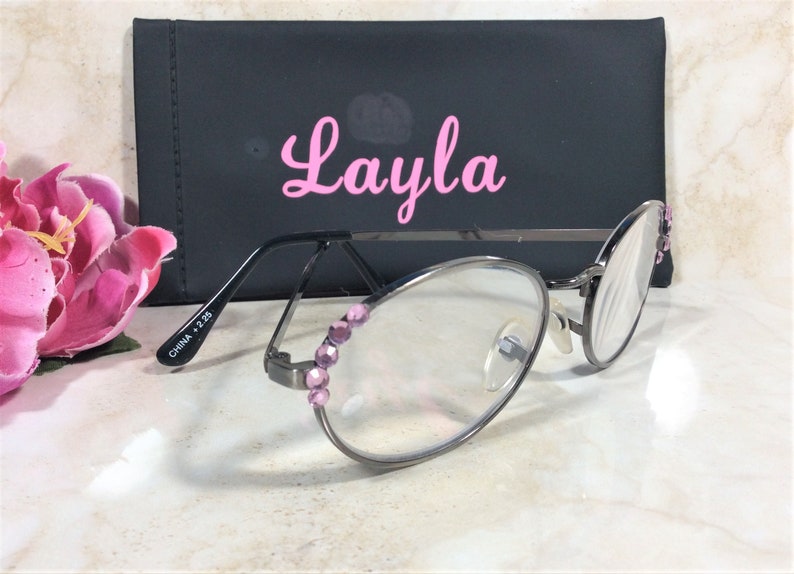 Puede incluir: Gafas con montura plateada, lentes transparentes y adornos de joyas rosas. Las gafas est&aacute;n frente a un estuche negro con el nombre "Layla" en escritura rosa. Una flor rosa est&aacute; en el fondo.