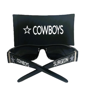 May include: Black sunglasses with the word "COWBOYS" and a star logo on the arms. The sunglasses are paired with a black case that has the word "COWBOYS" printed in white.