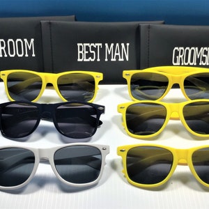 May include: A collection of sunglasses in yellow, black, and white frames with dark lenses, arranged in front of three black rectangular cases labeled with white text reading GROOM, BEST MAN, and GROOMSMAN in a bold, block-style font.