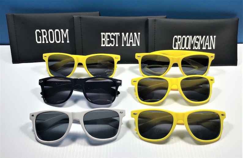 May include: A collection of sunglasses in yellow, black, and white frames with dark lenses, arranged in front of three black rectangular cases labeled with white text reading GROOM, BEST MAN, and GROOMSMAN in a bold, block-style font.