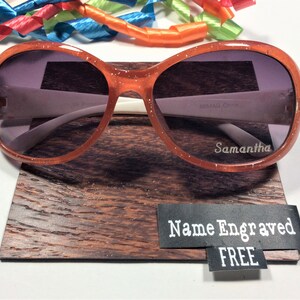 May include: Orange glitter sunglasses with a white frame and gray lenses. The sunglasses are on a wooden display stand with a black tag that says "Name Engraved FREE".