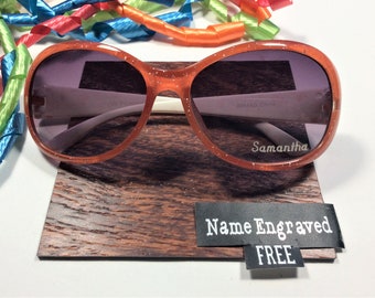 Puede incluir: Gafas de sol de color naranja con purpurina, con montura blanca y lentes grises. Las gafas de sol están sobre un expositor de madera con una etiqueta negra que dice "Nombre grabado GRATIS".