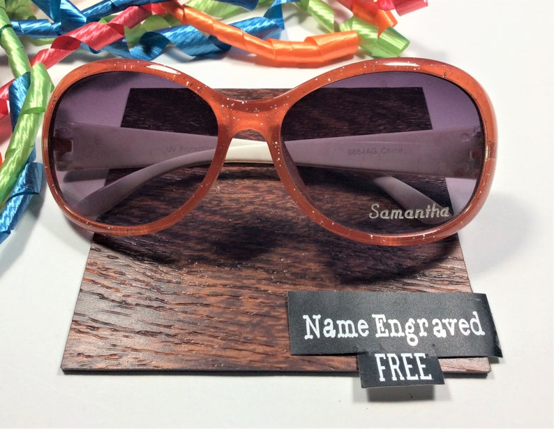 May include: Orange glitter sunglasses with a white frame and gray lenses. The sunglasses are on a wooden display stand with a black tag that says "Name Engraved FREE".