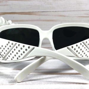 K&ouml;nnte beinhalten: Wei&szlig;e Sonnenbrille mit einem Nietendesign an den B&uuml;geln. Die Sonnenbrille hat schwarze Gl&auml;ser.