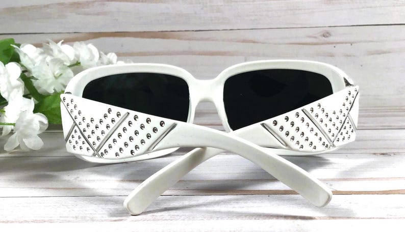 K&ouml;nnte beinhalten: Wei&szlig;e Sonnenbrille mit einem Nietendesign an den B&uuml;geln. Die Sonnenbrille hat schwarze Gl&auml;ser.
