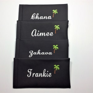May include: Four black eyeglass cases with white embroidered names and green palm tree designs. The names are "Chana", "Aimee", "Zahava", and "Frankie".