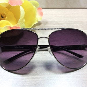 May include: Silver aviator sunglasses with dark purple lenses and rhinestones on the arms. The sunglasses have the text "TS2562-SLV" and "CHINA" printed on the arms.