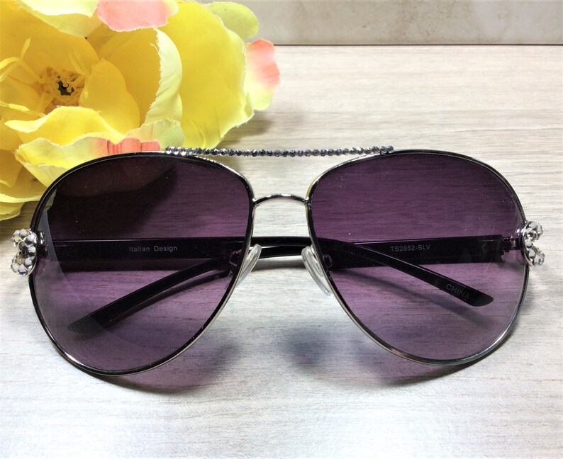 May include: Silver aviator sunglasses with dark purple lenses and rhinestones on the arms. The sunglasses have the text "TS2562-SLV" and "CHINA" printed on the arms.