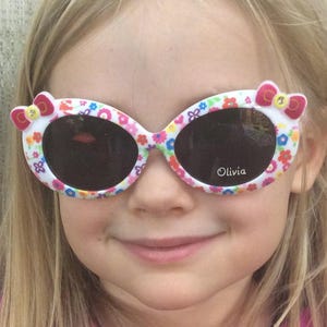 May include: A young girl wearing white sunglasses with a floral and pink bow design. The sunglasses have the word "Olivia" printed on the right lens.
