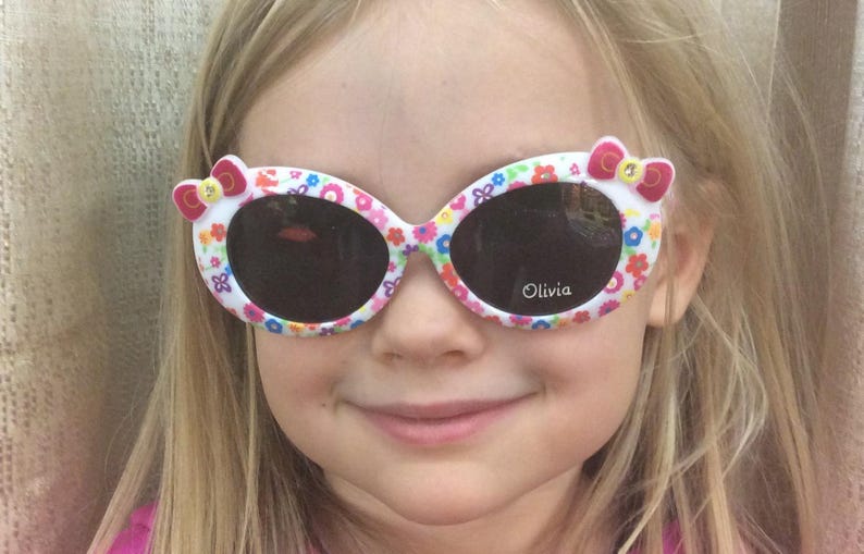 May include: A young girl wearing white sunglasses with a floral and pink bow design. The sunglasses have the word "Olivia" printed on the right lens.