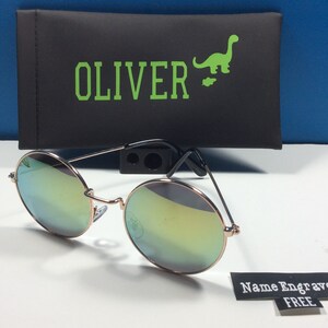 以下が含まれることがあります： 丸いミラーサングラス、ローズゴールドフレーム。サングラスは、ケースに「OLIVER」と緑色の恐竜のグラフィックが印刷された黒いケースに入っています。タグには「名前刻印無料」と書いてあります。