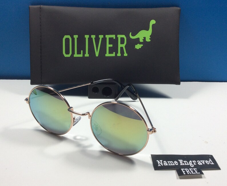 以下が含まれることがあります： 丸いミラーサングラス、ローズゴールドフレーム。サングラスは、ケースに「OLIVER」と緑色の恐竜のグラフィックが印刷された黒いケースに入っています。タグには「名前刻印無料」と書いてあります。