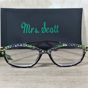 May include: Reading glasses with a dark frame, adorned with green and blue rhinestones along the top rim. The glasses are in front of a black case with the name "Mrs. Scott" in green script.