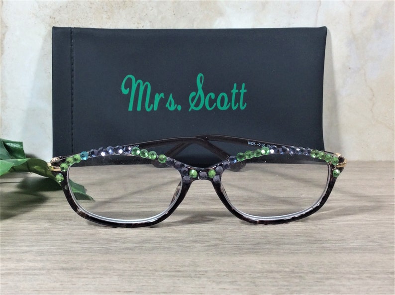 May include: Reading glasses with a dark frame, adorned with green and blue rhinestones along the top rim. The glasses are in front of a black case with the name "Mrs. Scott" in green script.