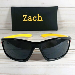 Op de afbeelding: Zwarte zonnebril met gele monturen en een rood logo aan de zijkant. De zonnebril ligt op een wit houten oppervlak. Een zwarte etui met de naam "Zach" erop geborduurd is op de achtergrond.