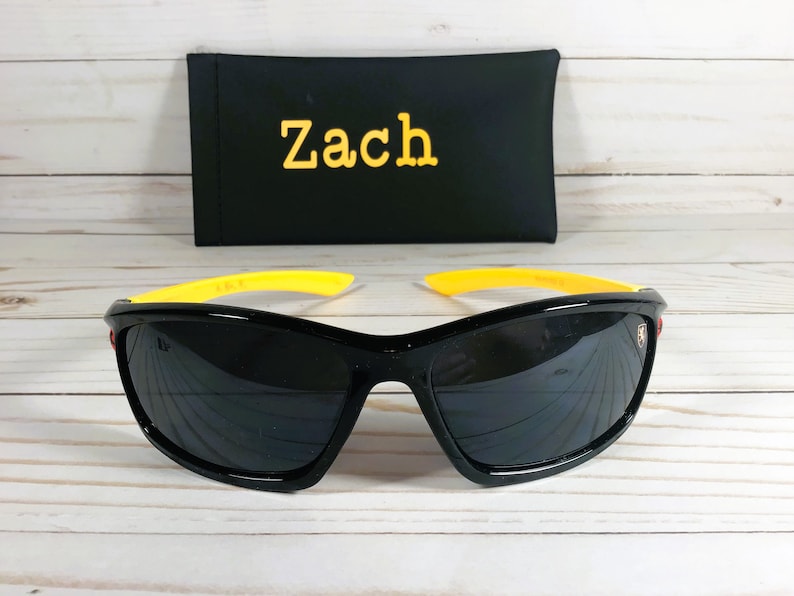 Op de afbeelding: Zwarte zonnebril met gele monturen en een rood logo aan de zijkant. De zonnebril ligt op een wit houten oppervlak. Een zwarte etui met de naam "Zach" erop geborduurd is op de achtergrond.