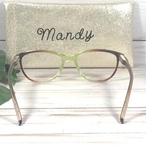 Pu&ograve; includere: Un paio di occhiali con montatura sfumata marrone e verde, davanti a un astuccio dorato. L'astuccio presenta il nome "Mandy" scritto in corsivo.
