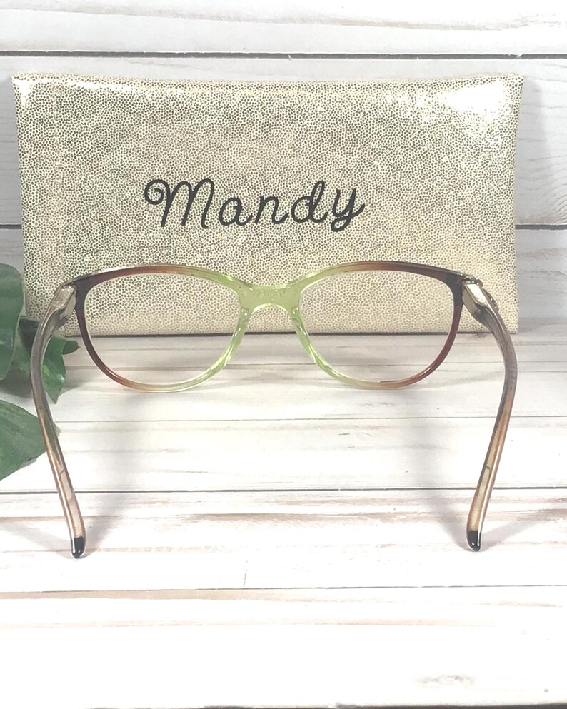 Pu&ograve; includere: Un paio di occhiali con montatura sfumata marrone e verde, davanti a un astuccio dorato. L'astuccio presenta il nome "Mandy" scritto in corsivo.