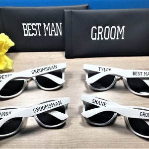 May include: Set of six white sunglasses with black lenses. Each pair has a different name printed on the frame: Pete, Danny, Groom, Shane, Tyler, and Best Man. The sunglasses are displayed on a wooden surface with two black cases labelled "Best Man" and "Groom".