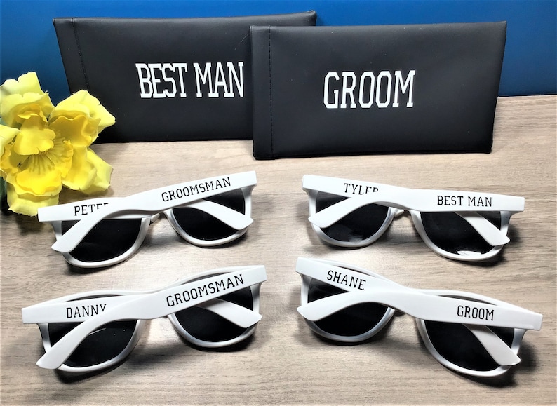 May include: Set of six white sunglasses with black lenses. Each pair has a different name printed on the frame: Pete, Danny, Groom, Shane, Tyler, and Best Man. The sunglasses are displayed on a wooden surface with two black cases labelled "Best Man" and "Groom".