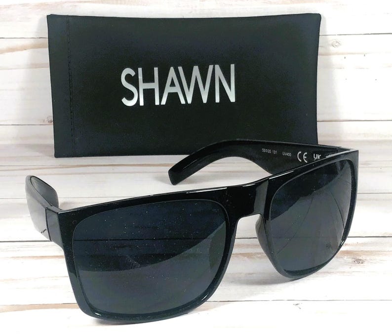 Puede incluir: Gafas de sol negras con montura rectangular y lentes oscuras. Las gafas de sol est&aacute;n junto a un estuche negro con el nombre "SHAWN" impreso en plata. Las gafas tienen una marca CE y UK.