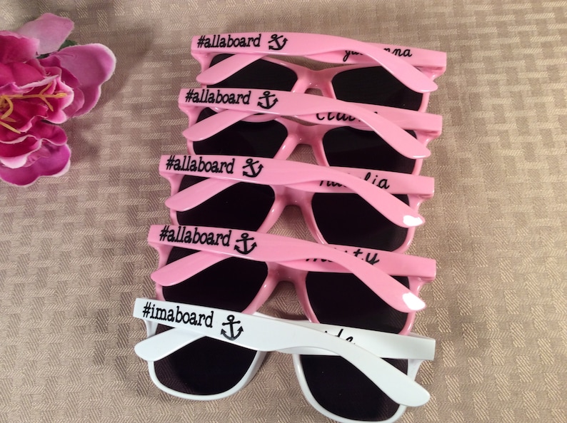 May include: Five pink sunglasses with black lenses and a black anchor and the text "#allaboard" printed on the arms. One white pair of sunglasses with black lenses and a black anchor and the text "#imaboard" printed on the arms.