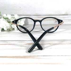 May include: Black spectacles with a rounded frame shape and thin metallic gold-toned temples. The arms of the glasses are folded inwards, crossing each other at the centre. The frame features a glossy finish and clear lenses, showcasing a sleek design.