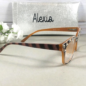 Puede incluir: Gafas marrones y beige con adornos de pedrer&iacute;a. Las monturas son de color marr&oacute;n claro y las patillas tienen un dise&ntilde;o a rayas. En el fondo, una funda plateada con el nombre "Alexia".