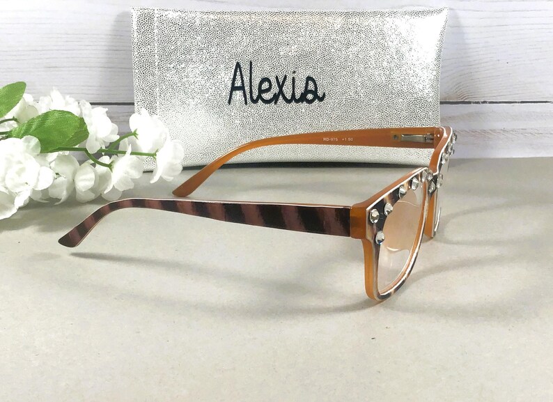 Puede incluir: Gafas marrones y beige con adornos de pedrer&iacute;a. Las monturas son de color marr&oacute;n claro y las patillas tienen un dise&ntilde;o a rayas. En el fondo, una funda plateada con el nombre "Alexia".