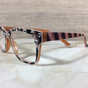 Puede incluir: Gafas con montura marr&oacute;n y estampado de cebra en las patillas. Las monturas est&aacute;n adornadas con brillantes transparentes. Las lentes son transparentes y las gafas est&aacute;n sobre un fondo neutro.
