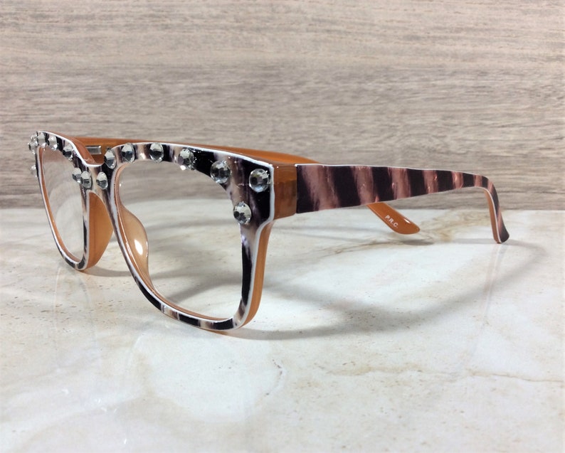 Puede incluir: Gafas con montura marr&oacute;n y estampado de cebra en las patillas. Las monturas est&aacute;n adornadas con brillantes transparentes. Las lentes son transparentes y las gafas est&aacute;n sobre un fondo neutro.
