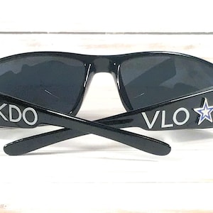 May include: Black sunglasses with dark lenses. The temples feature the letters "KDO" and "VLO" in white, along with a white star outline with a blue center. The sunglasses are set against a white wooden background.