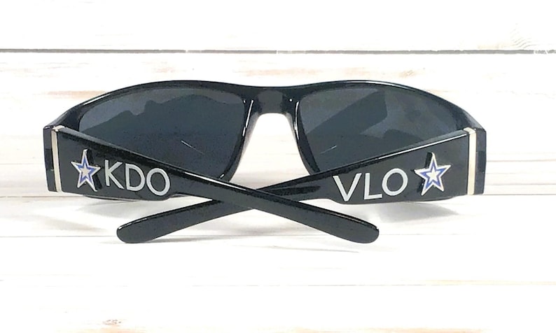 May include: Black sunglasses with dark lenses. The temples feature the letters "KDO" and "VLO" in white, along with a white star outline with a blue center. The sunglasses are set against a white wooden background.