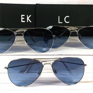 May include: Silver-framed aviator sunglasses with gradient blue lenses. The sunglasses are displayed with two black cases, each with white initials. One pair has "EK" and "LC" on the lenses.