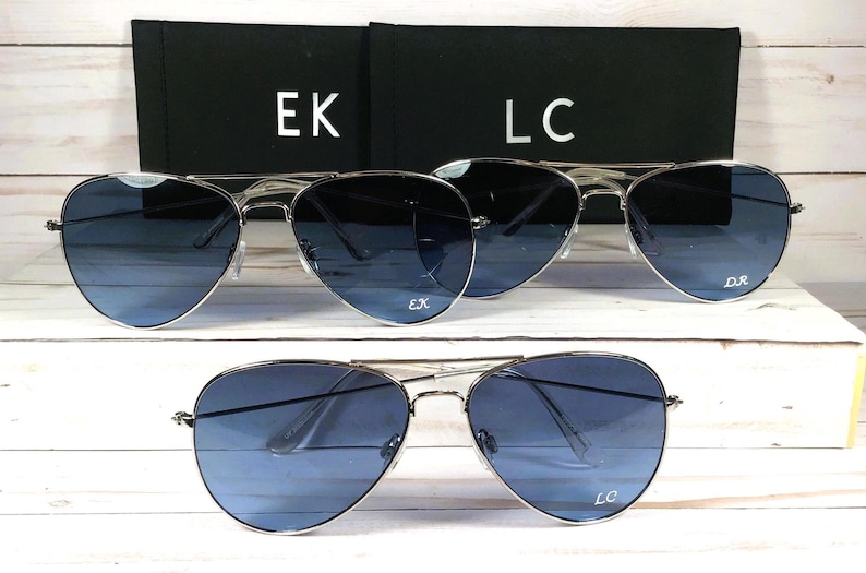 May include: Silver-framed aviator sunglasses with gradient blue lenses. The sunglasses are displayed with two black cases, each with white initials. One pair has "EK" and "LC" on the lenses.