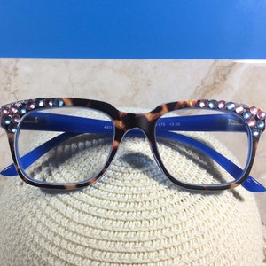 May include: Eyeglasses with a tortoiseshell pattern frame and blue arms. The top of the frame is embellished with pink and blue rhinestones. The glasses are resting on a white hat.