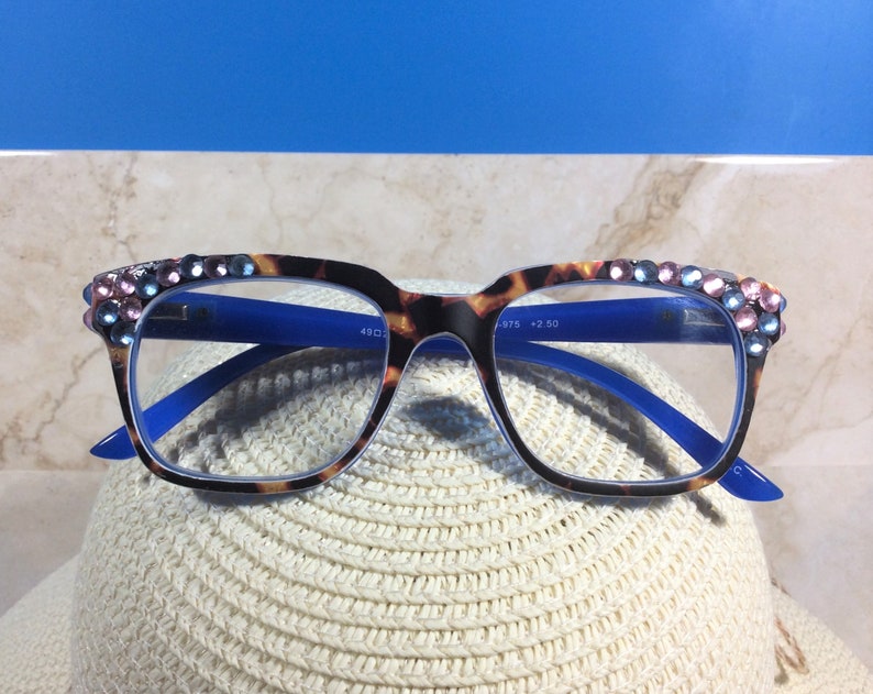 May include: Eyeglasses with a tortoiseshell pattern frame and blue arms. The top of the frame is embellished with pink and blue rhinestones. The glasses are resting on a white hat.