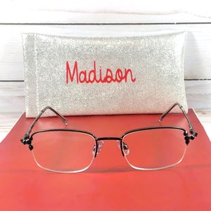 May include: A pair of eyeglasses with a black frame and clear lenses, resting on a red surface. The glasses have decorative accents on the temples. Behind the glasses is a silver case with the name "Madison" embroidered in red.