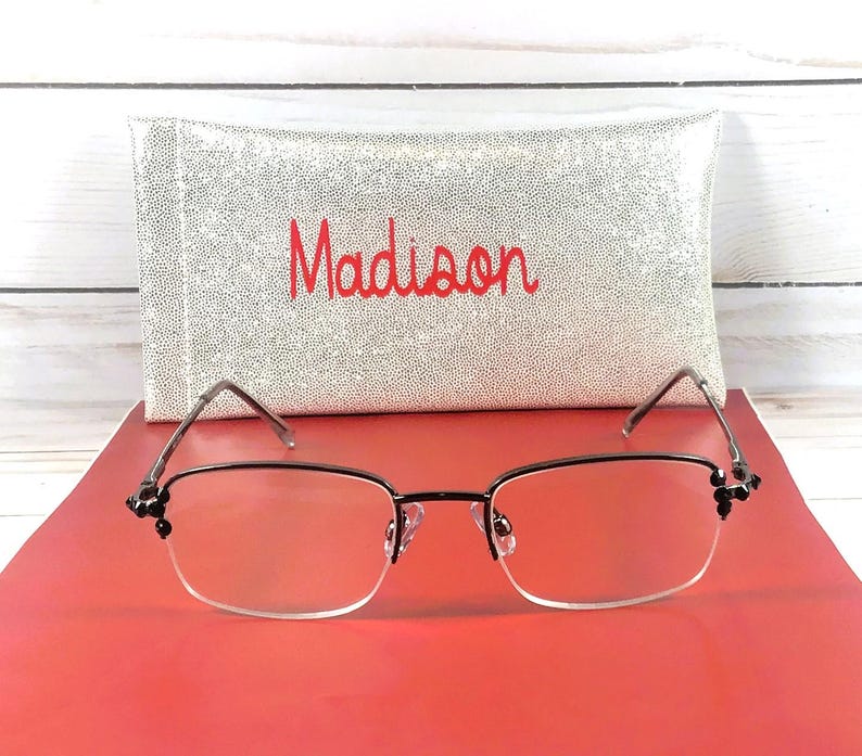 May include: A pair of eyeglasses with a black frame and clear lenses, resting on a red surface. The glasses have decorative accents on the temples. Behind the glasses is a silver case with the name "Madison" embroidered in red.