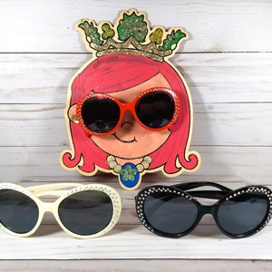 May include: Three pairs of sunglasses with rhinestones on the frames. The sunglasses are white, black, and red. The red sunglasses are on a painted wooden cutout of a girl with pink hair wearing a crown.