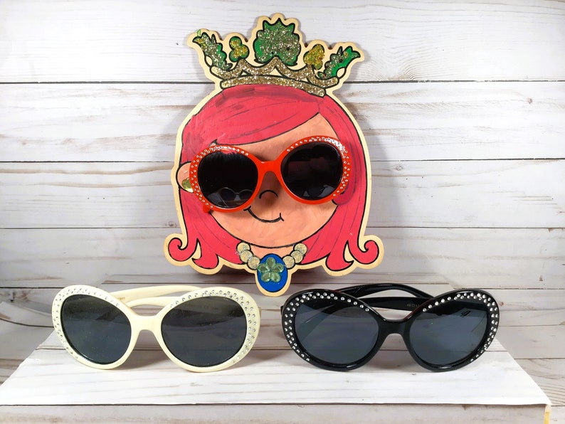 May include: Three pairs of sunglasses with rhinestones on the frames. The sunglasses are white, black, and red. The red sunglasses are on a painted wooden cutout of a girl with pink hair wearing a crown.
