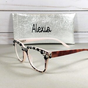 May include: A pair of eyeglasses with a light pink frame, embellished with black and clear rhinestones along the top edge. The temples are a brown tortoiseshell pattern. A silver case with the name "Alexia" is behind the glasses.
