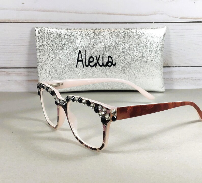 May include: A pair of eyeglasses with a light pink frame, embellished with black and clear rhinestones along the top edge. The temples are a brown tortoiseshell pattern. A silver case with the name "Alexia" is behind the glasses.