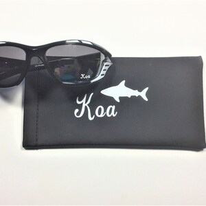 May include: Black sunglasses with white accents and a black case with a white shark and the word "Kao" printed on it.