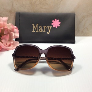 May include: Brown and gold-toned sunglasses with a chain detail on the frame. The sunglasses are in a black case with the name "Mary" and a pink flower printed on it.