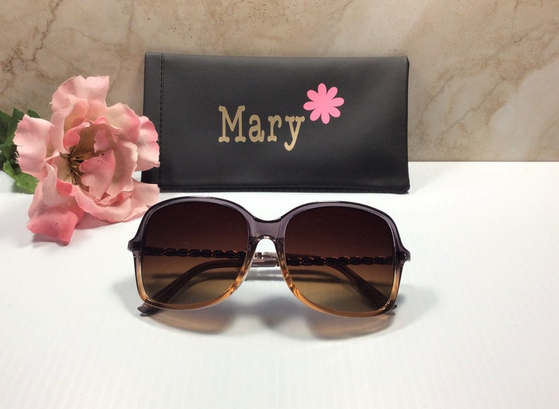 May include: Brown and gold-toned sunglasses with a chain detail on the frame. The sunglasses are in a black case with the name "Mary" and a pink flower printed on it.