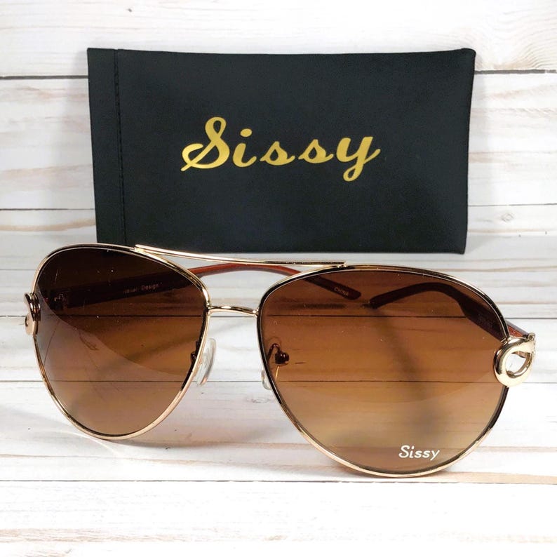 May include: Gold-rimmed aviator sunglasses with brown gradient lenses. The sunglasses have a double bridge and a decorative detail on the temples. A black case with the word "Sissy" in gold script is behind the sunglasses.