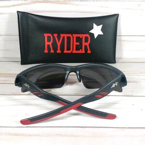 K&ouml;nnte beinhalten: Schwarze Sonnenbrille mit roten Akzenten und einem goldenen Logo. Die Sonnenbrille ist zusammengeklappt und liegt auf einer Holzoberfl&auml;che. Die Sonnenbrille wird mit einem schwarzen Etui geliefert, auf dem der Name "RYDER" und ein wei&szlig;er Stern aufgedruckt sind.