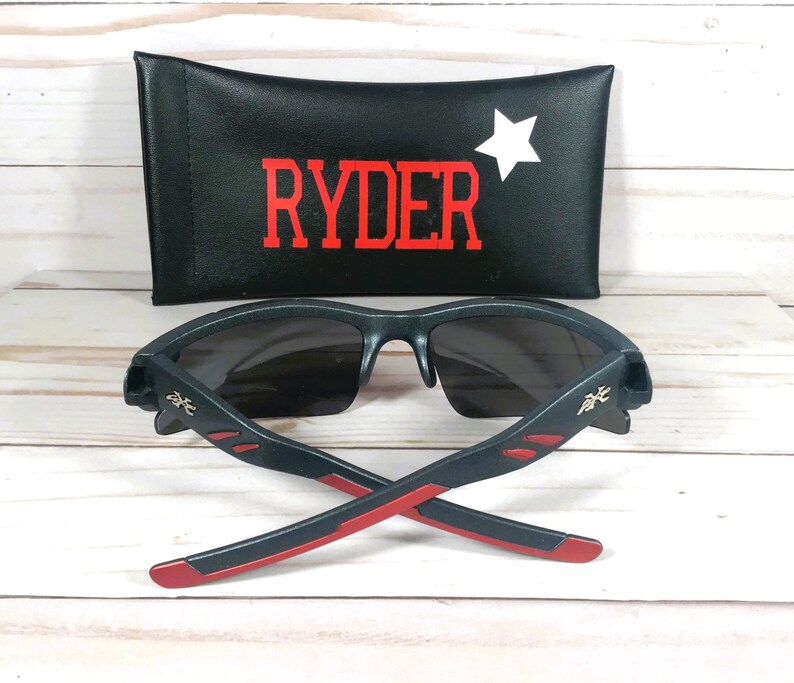 K&ouml;nnte beinhalten: Schwarze Sonnenbrille mit roten Akzenten und einem goldenen Logo. Die Sonnenbrille ist zusammengeklappt und liegt auf einer Holzoberfl&auml;che. Die Sonnenbrille wird mit einem schwarzen Etui geliefert, auf dem der Name "RYDER" und ein wei&szlig;er Stern aufgedruckt sind.