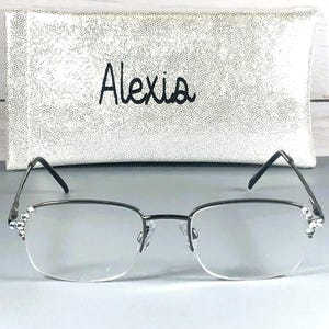 Pode incluir: &Oacute;culos com arma&ccedil;&atilde;o prateada e lentes transparentes, com strass decorativos nas hastes. Atr&aacute;s dos &oacute;culos, uma capa de &oacute;culos prateada e texturizada com o nome "Alexis" bordado em preto. A capa tem um padr&atilde;o de pontos.